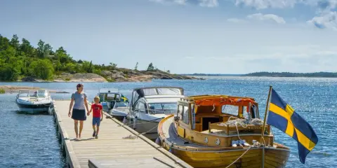 En kvinna och ett barn går på en brygga med båtar och en svensk flagga. En perfekt bild av en semester i Sverige.
