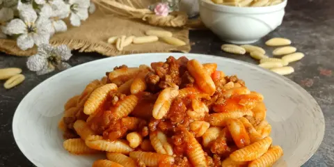 Un piatto invitante di malloreddus alla campidanese, conditi con un ricco ragù di carne. Sullo sfondo, fiori bianchi e un ciotola di pasta cruda.
