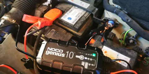 Ładowarka NOCO Genius 10 podłączona do akumulatora samochodowego, który potrzebuje ładowania. Czerwone kable z zaciskami podłączone do biegunów.