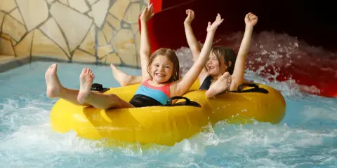 Zwei lachende Kinder rutschen auf einem gelben Schlauchboot durch einen Wasserpark. Ein perfekter Tag, wie ein h2o gutschein!