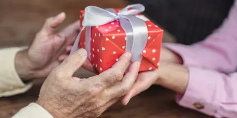 Dłonie przekazują sobie prezent w czerwonym papierze w białe kropki z białą wstążką. To piękny gest z okazji 14 rocznicy ślubu.