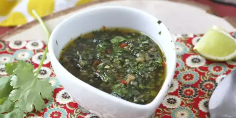Aromaticzny sos chimichurri w białej miseczce, obok ćwiartka limonki i świeża kolendra. Idealny do grillowanych mięs.