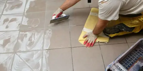 Pracownik w rękawiczkach nakłada fugę epoksydową na płytki podłogowe za pomocą kielni. Proces fugowania krok po kroku.