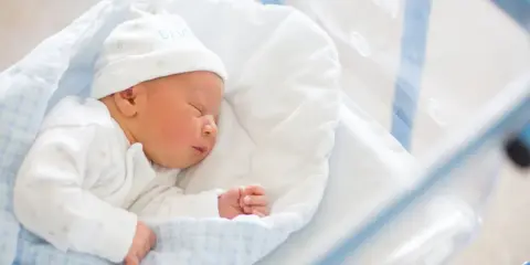 Śpiący noworodek w szpitalnym łóżeczku. Do wniosku o becikowe potrzebne są dokumenty potwierdzające urodzenie dziecka.