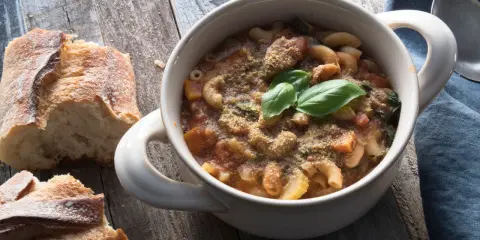 Una ciotola di pasta e fagioli in scatola, guarnita con basilico fresco e pane croccante su un tavolo di legno.