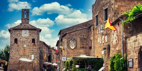 Un vicolo pittoresco a Sovana, Italia, con edifici in pietra, bandiere colorate e un cielo azzurro pieno di nuvole.