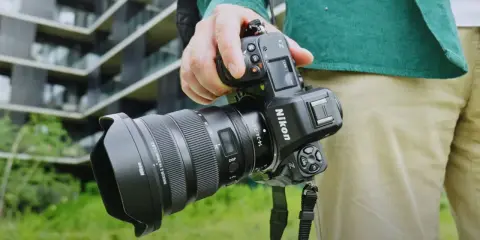 Dłoń trzyma aparat Nikon Z8 z obiektywem. W tle nowoczesny budynek.