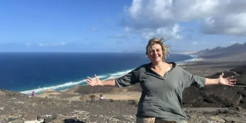 Donna sorridente con le braccia aperte su un paesaggio costiero, un'espressione di gioia che contrasta la solitudine interiore.