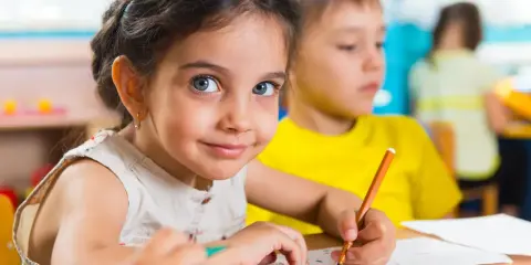 Uśmiechnięta dziewczynka z niebieskimi oczami rysuje kredką. Jej przeniesienie dziecka do innego przedszkola nie zasmuciło jej.