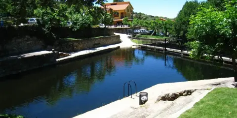 La piscina natural de Casas del Monte, rodeada de vegetación y con un edificio al fondo, invita a un refrescante baño.