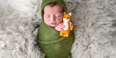 Dolci immagini neonati: un bimbo avvolto in una coperta verde tiene stretto un piccolo volpino di peluche, dormendo sereno su un soffice tappeto bianco.