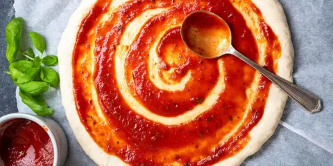 Pizzaboden mit selbstgemachter Pizzasauce bestreichen. Frisches Basilikum und ein Löffel mit Sauce daneben.