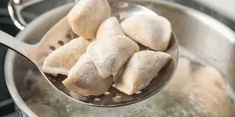 Mrożone pierogi na łyżce cedzakowej nad gotującą się wodą.