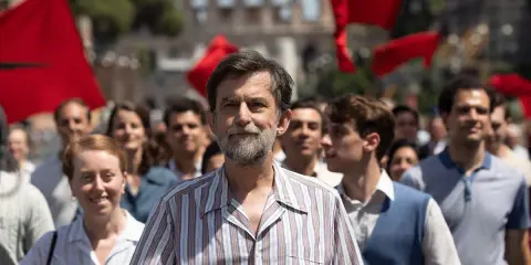 Nanni Moretti cammina in una folla con bandiere rosse, sullo sfondo il Colosseo.