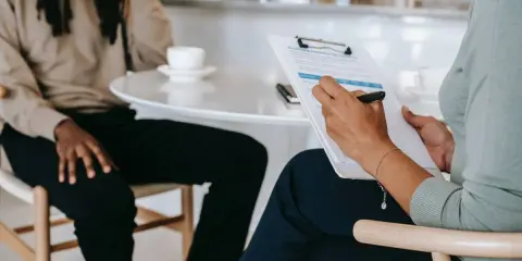 Osoba z dredami siedzi przy stole, podczas gdy druga osoba zapisuje odpowiedzi na pytania rekrutacyjne po angielsku.