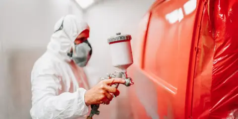 Pracownik w kombinezonie ochronnym maluje maskę samochodu na jaskrawy czerwony kolor.