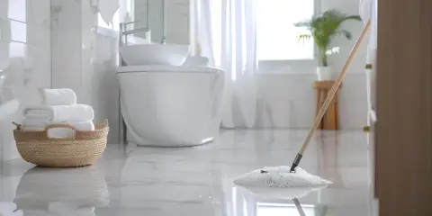 Czyszczenie łazienki w toku. Mop delikatnie przesuwa się po lśniącej podłodze, a stos białych ręczników czeka w wiklinowym koszu.