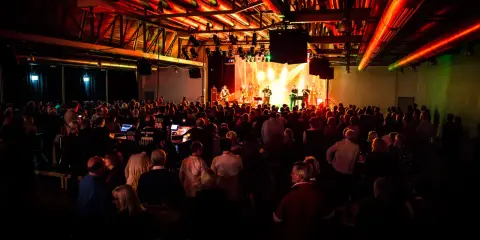 Ein Konzert in Emsdetten mit einer Band auf der Bühne und vielen begeisterten Zuschauern. Die Beleuchtung taucht die Szene in ein warmes, rotes Licht.