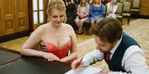 Panna młoda w czerwonej sukni i pan młody podpisują dokumenty. W tle goście robią zdjęcia telefonami. Może to być ślub, gdzie nie mam świadków.