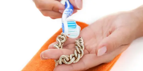 Manos limpian una pulsera de oro con un cepillo de dientes y un paño naranja. Aprende como limpiar el oro para que brille.