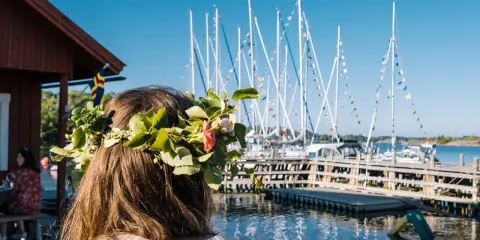 En flicka med blomsterkrans ser ut över en hamn med segelbåtar. En perfekt dag för en resa till Åland.