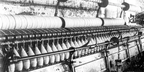Macchinario industriale per la filatura con fili avvolti su bobine e grandi rulli.