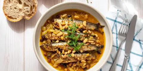 Sarde in saor in una ciotola, con pinoli, uvetta e cipolle. Accanto, pane rustico e posate.