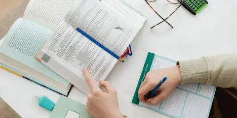 Ręce studenta nad otwartymi książkami, notatnik z napisem "I WILL BE A GREAT STUDENT", okulary i kalkulator. Nauka do egzaminu, by zdobyć tytuł zawodowy.