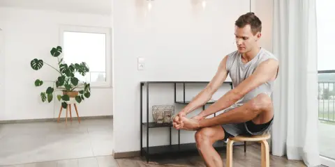 Mann sitzt auf einem Hocker und dehnt seine Zehen. Ein Monstera-Pflanze steht im Hintergrund.