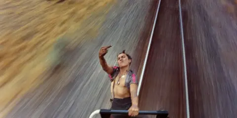 Mike Brodie, un giovane ribelle, fa il gesto del dito medio mentre viaggia su un treno merci, sfidando il mondo con uno sguardo provocatorio.