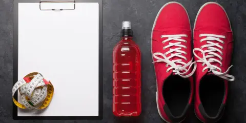 Tabela węglowodanów w produktach, miarka krawiecka, czerwony napój izotoniczny i buty sportowe.