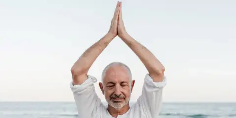 Mężczyzna z zamkniętymi oczami, ręce złożone nad głową, praktykuje mindfulness co to. Spokój nad morzem.