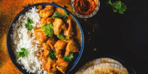 Aksamitny kurczak w sosie butter masala, podany z ryżem i chlebkami naan. Obok miseczka z przyprawami.