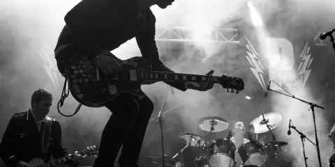 Un guitariste électrique joue sur scène, entouré de fumée. Derrière lui, un batteur et le logo d'un nom de groupe de rock.