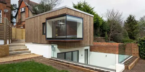 Une extension de maison moderne en bois avec une grande fenêtre en saillie. L'agrandissement maison s'intègre à un jardin en pente avec des murs de briques et des garde-corps en verre.
