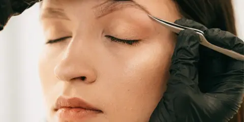 Une main gantée retire délicatement un poil incarné au sourcil d'une femme aux yeux fermés.