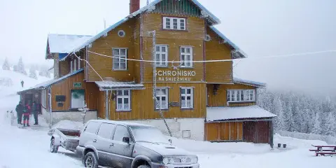 Schronisko na Śnieżniku w zimowej scenerii. Samochód z przyczepą i turyści przy wejściu. Masyw Śnieżnika i jego szlaki w śniegu.