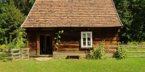 Drewniana chata z glinianym dachem, typowa dla skansenów w Polsce, otoczona zielenią i drewnianym płotem.