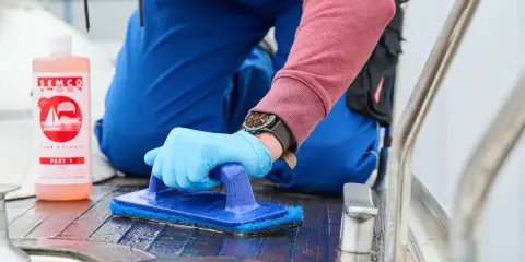 Person reinigt mit blauer Bürste und SEMCO Teak Cleaner das Holzdeck eines Bootes.