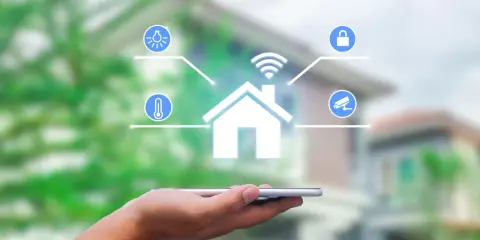 Dłoń trzyma smartfon, z którego wyłania się symbol domu z ikonami oświetlenia, temperatury, zamka i kamery. To wizualizacja systemu zarządzania energią.