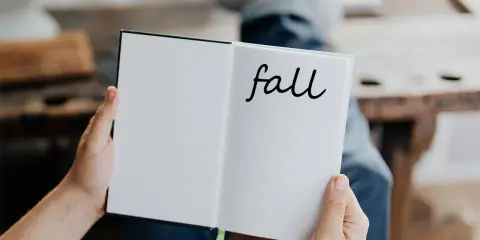 Otwarta książka z napisem "fall" na prawej stronie. Osoba w dżinsach trzyma ją, przygotowując się do jesiennych zapisków.