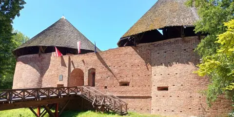 Zamek międzyrzecz z drewnianym mostem i wieżami o stożkowych dachach. Czerwona cegła i zieleń otoczenia.