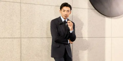 A man in a black suit and black shirt, holding sunglasses, leans against a textured wall.