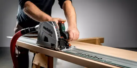 Main d'un artisan guidant une scie circulaire sur un rail pour une coupe précise sur une planche de bois.
