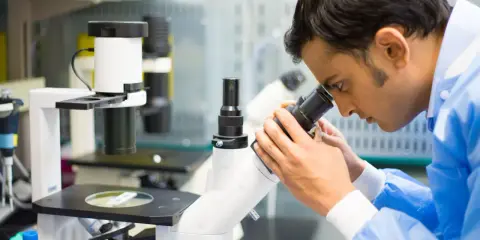 Mężczyzna w niebieskim fartuchu laboratoryjnym patrzy przez mikroskop.
