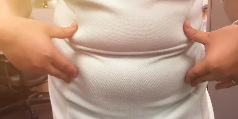 Dłonie zaciskają białą tkaninę, uwydatniając zaokrąglony brzuch. Może to być objaw problemów z tarczycą, czyli tzw. brzuch tarczycowy.