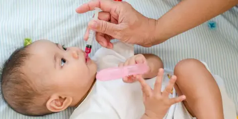 Dłoń podaje niemowlęciu witaminę D z pipetki. Dziecko leży na boku, ubrana w białą koszulkę.