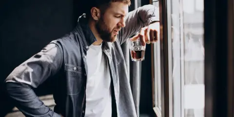 Mężczyzna zamyślony przy oknie, trzymający szklankę z bursztynowym płynem. Czy to jeden z ukryty alkoholizm objawy?