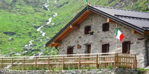 Rifugio in pietra con bandiera italiana, circondato da verdi pendii e un ruscello. Un'oasi accogliente tra i rifugi Val Camonica facili da raggiungere.
