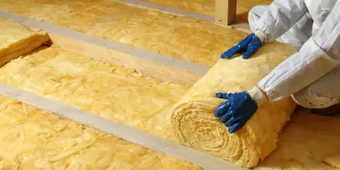 Pose d'isolant jaune, peut-être laine de verre vs laine de roche, dans un grenier. Mains gantées bleues déroulant un rouleau.
